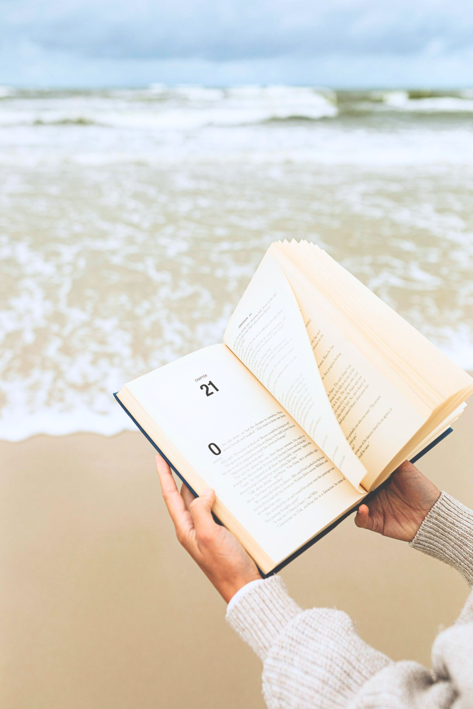 hands holding a book open with pages blowing in the wind at the beach with the shoreline in the background