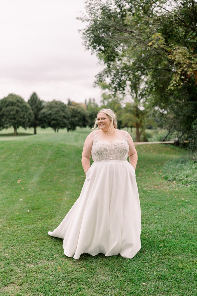 Shay standing on grass in a wedding dress with hands in her pockets and trees in the background
